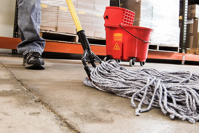 Worker cleaning a warehouse floor with a mop beside a red cleaning bucket